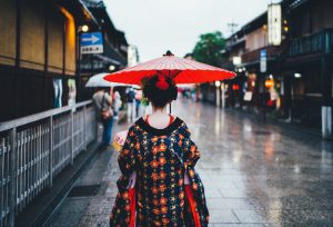 Picture presents geisha walking on a rainy streets of Kyoto. She is wearing a traditional kimono and holding a red umbrella.